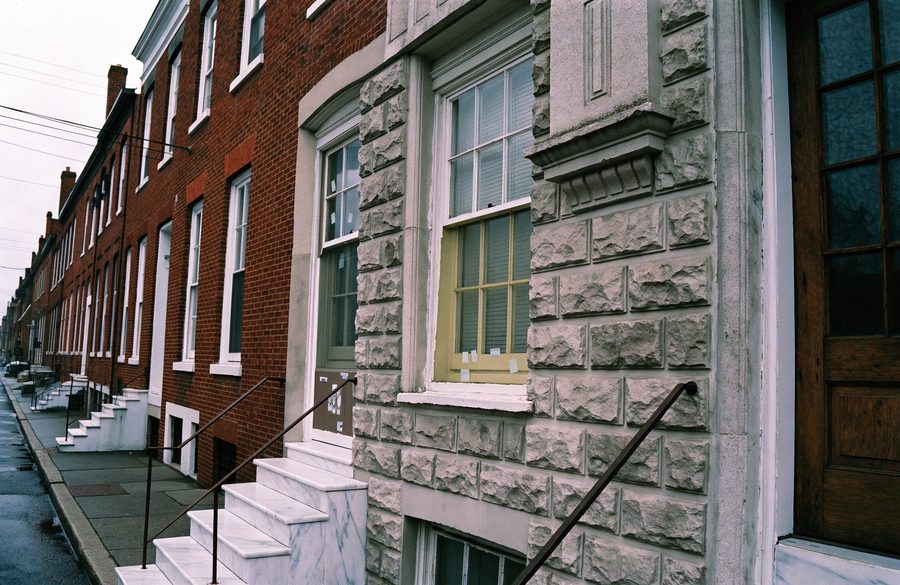A Formstone-clad Baltimore rowhome beside plain brick neighbours, white marble steps at the pavement and brick chimney stacks above
